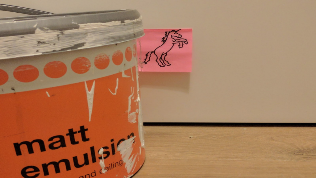 Same unicorn post-it as in previous photo. The rainbow lamp is gone, replaced by a large tub of matt emulsion. It looks like a cheap brand of paint, and the tub has messy paint spodges and some cobwebs on it.