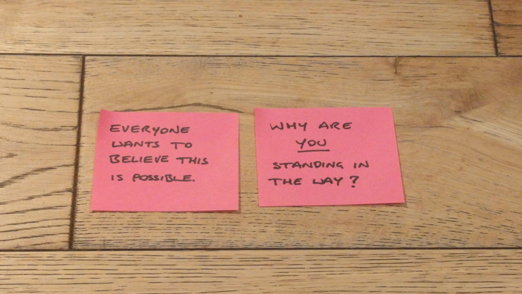 Post-its on a wooden floor:

"Everyone wants to believe this is possible"
"Why are you standing in the way?"

In the second post-it, "you" is underlined.