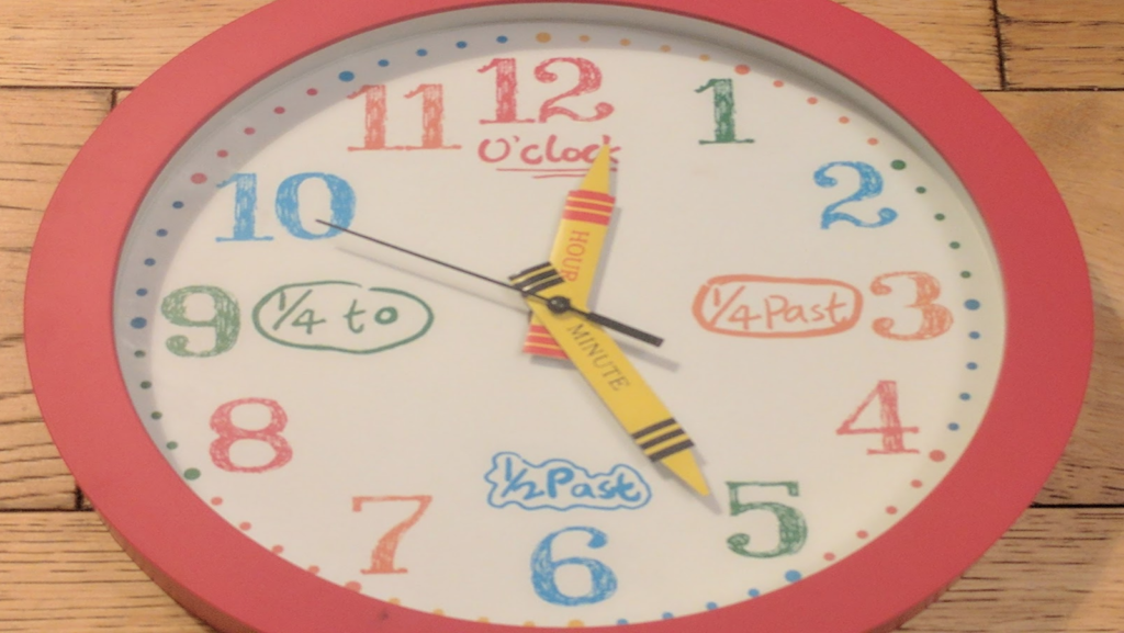 Clock lying on the wooden floor. The clock is made for a kid's room: The hands are crayons (with "hour" and "minute" where the crayon colour is usually written) and the numbers round the outside look like they've been drawn with crayons.