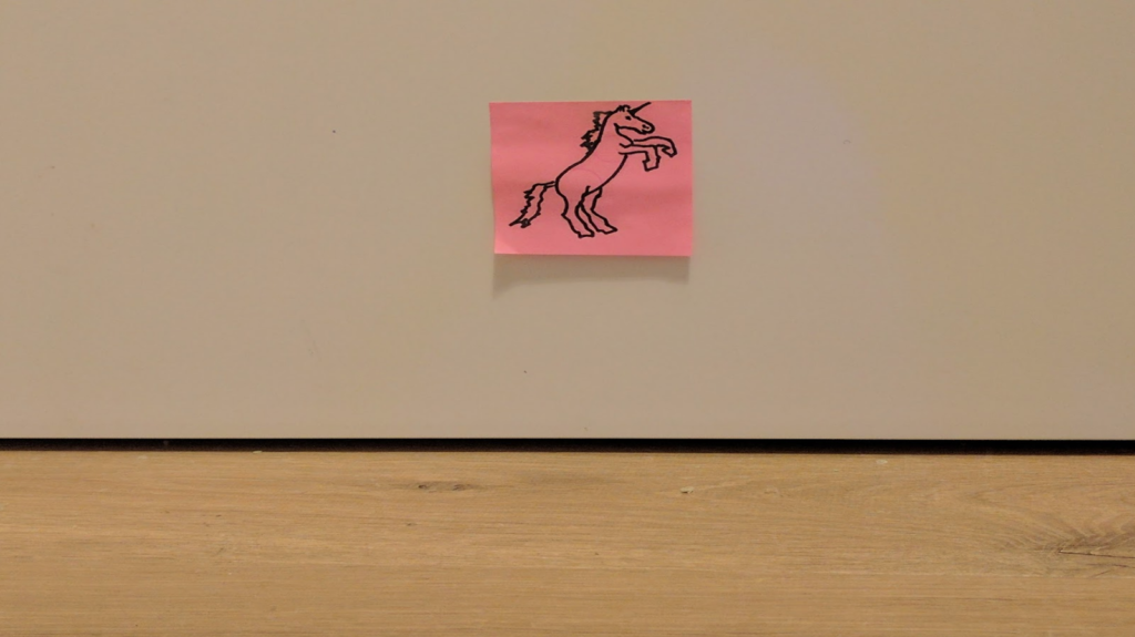 Photo of a post-it with a unicorn drawn on it, rearing up on its back hooves. The post-it is stuck to a cupboard above a wooden floor.