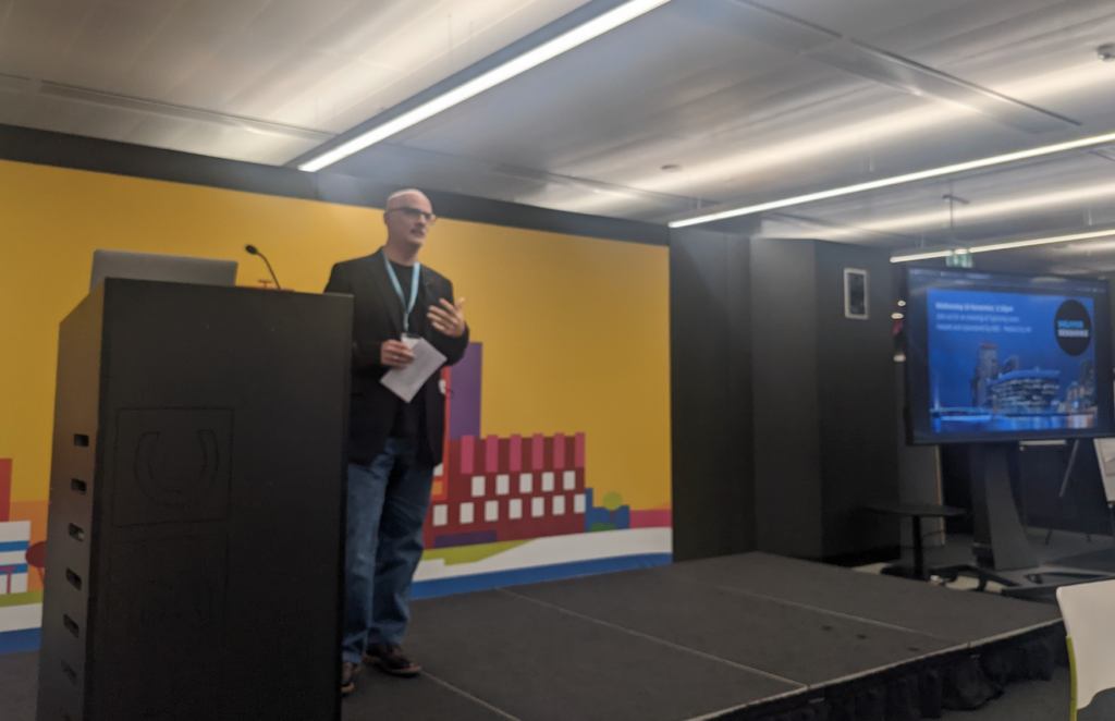 Speaker on stage next to a lectern. The stage has a colourful background. To the left, the poster for the event (from the top of this blog post) is showing on a large TV screen.