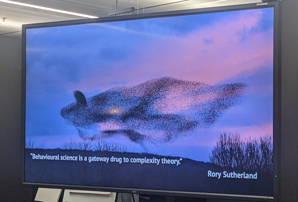 Side with a photo of a huge flock of birds making a pattern. Quote at the bottom: "Behavioural science is a gateway drug to complexity theory" (Rory Sutherland).