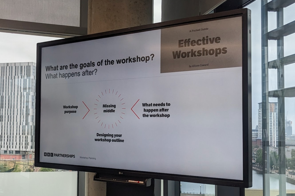 Photo of a slide on a big screen, showing the "Effective Workshops book"

"Whate are the goals of the workshop? What happens after?"

Diagram shows "workshop purpose" pointing in from the left, and "what needs to happen after the workshop" from the right, with a dashed circle saying "Missing middle" between them.

Under the middle part: "developing your workshop outline"