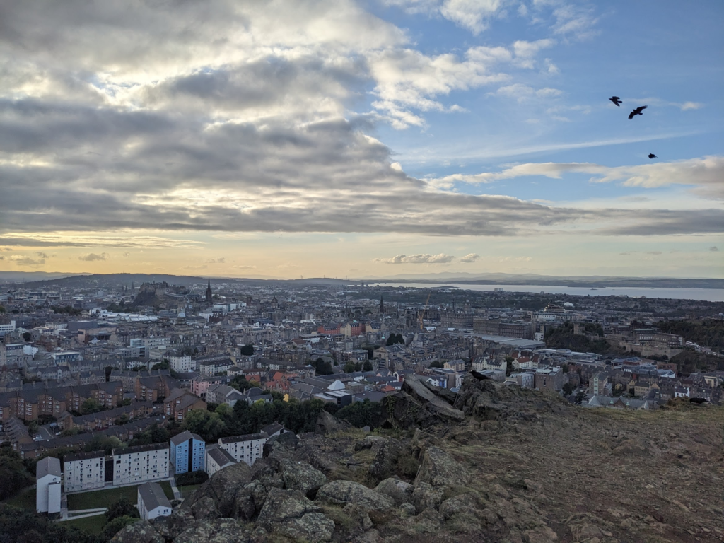 Photo from high above Edinburgh, looking across the city out to the Firth of Forth. Crows fly in a sunny sky with dramatic clouds.