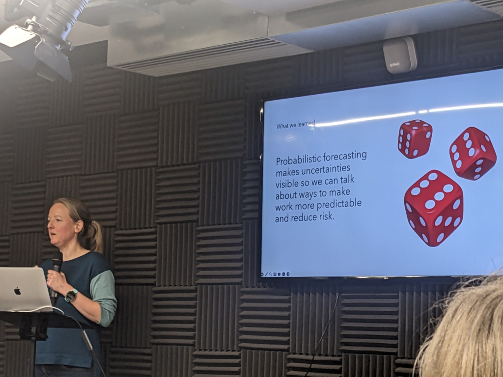 Sarah at the front of the room with a microphone, with a big screen behind her. Screen has an image of 3 dice, and words: "Probabilistic forecasting makes uncertainties visible so we can talk about ways to make work more predictable and reduce risk"