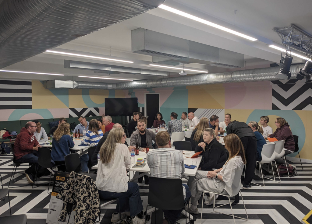 Tables of people discussing the talk - about 30-something people in total. The room is colourfully decorated, with zigzag lines on the floor and large circles and triangles on the walls.