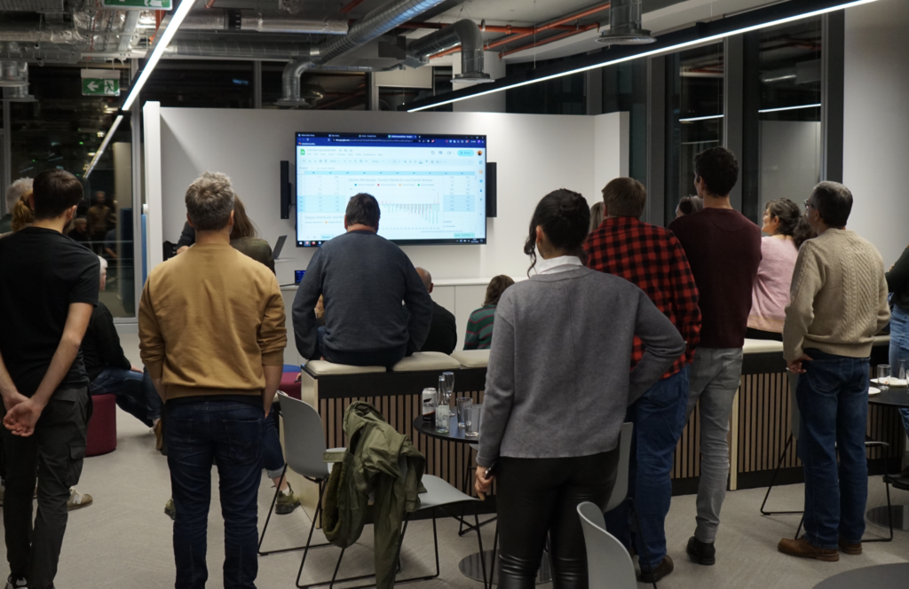 Photo of the attendees at Deliver Sessions: everyone's stood up (waiting to go and sit with their teams), looking at the big screen where Mark is explaining the game.