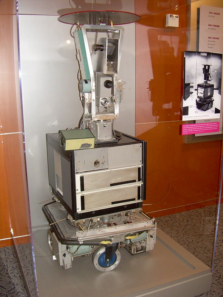 Colour photo of Shakey the robot in a glass display case. On the front of the case, at eye level, is the same labelled 1972 photo of Shakey that was included at the start of this blog post.