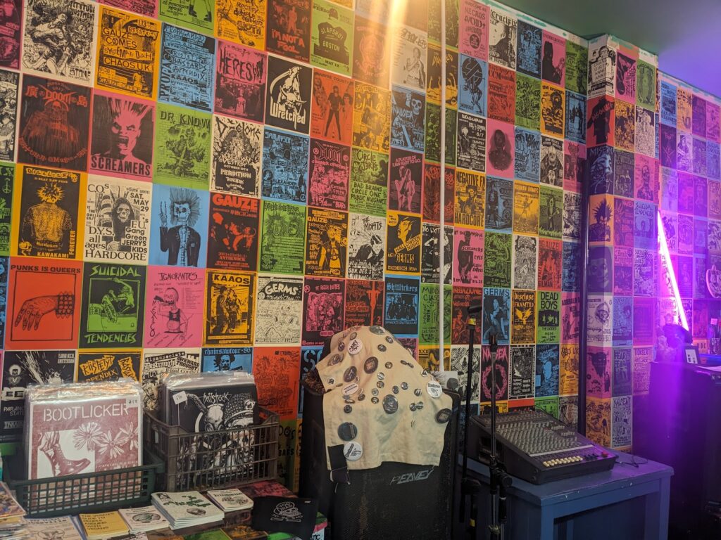 Photo of a wall decorated with band posters, with racks of vinyl records in front of it. There's also a microphone stand and a mixing desk.