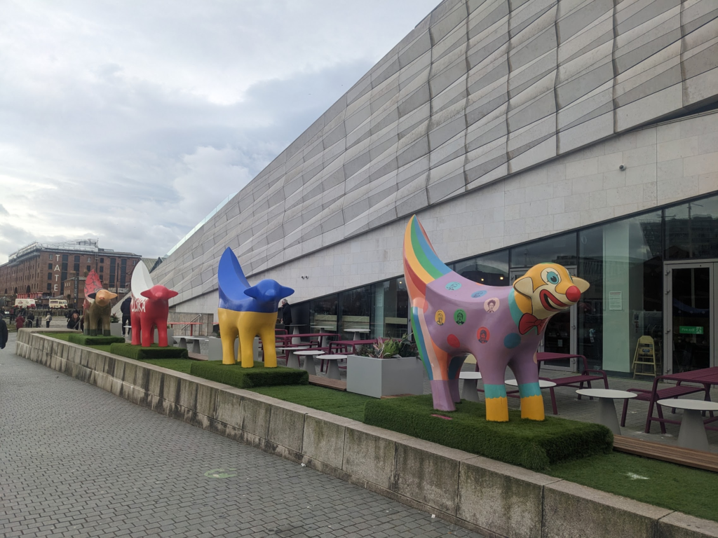 A line of brightly painted sculptures in front of a museum. Each sculpture is the same shape: a lamb/banana combination.