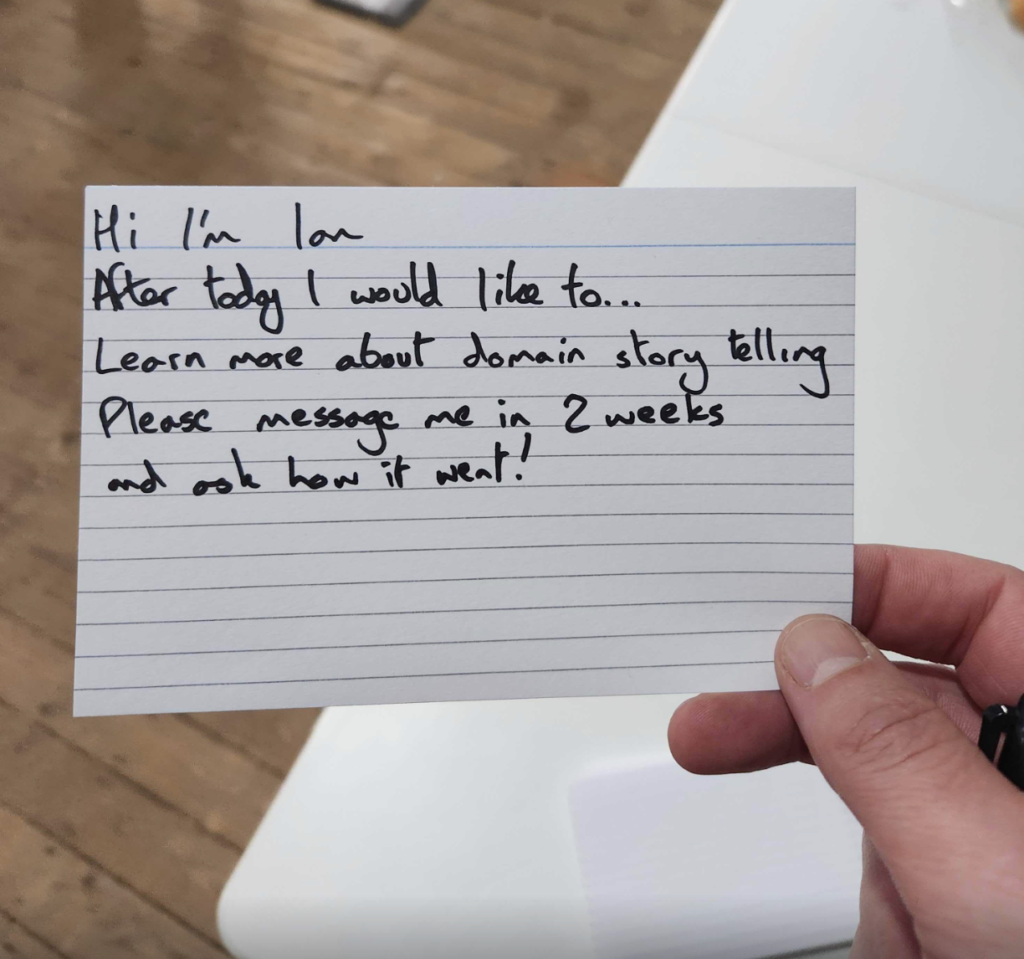 Photo of a hand holding an index card. Card says "Hi I'm Ian. After today I would like to... learn more about domain storytelling. Please message me in 2 weeks and ask how it went!"