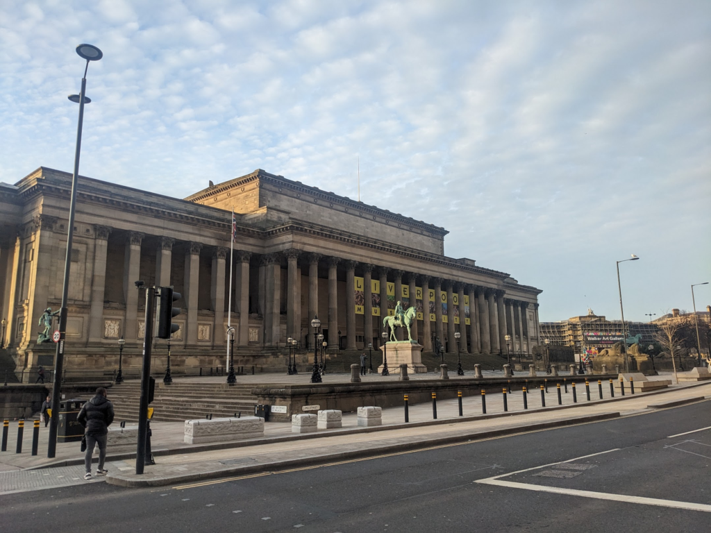 Photo of a large building with a long row of columns, and statues in front of it. Sky is blue with fluffy clouds.