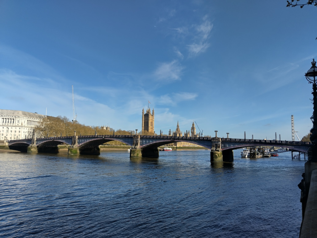 Photo of a sunny blue sky and blue river, with a bridge and Houses of Parliament in between.