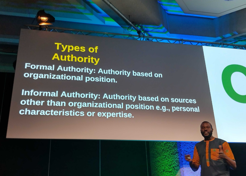 Photo of a slide on a huge screen, with the speaker (Eb) in front of it. Slide title is "Types of authority", and says:

"Formal authority: authority based on organizational position.

Informal authority: authority based on sources other than organizational position e.g., personal characteristics or expertise."