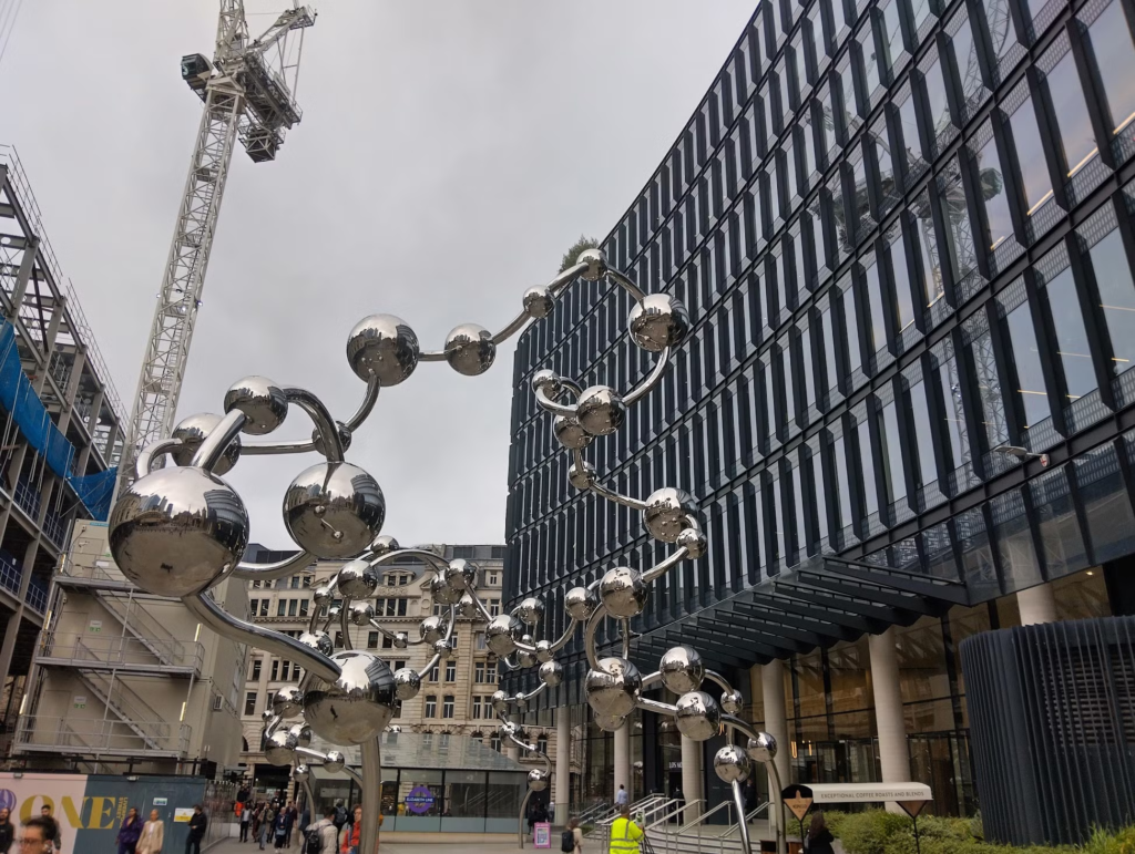 Photo of London, with buildings on other side of a dramatic sculpture: lots of reflective metal balls spaced along a twisting metal support.