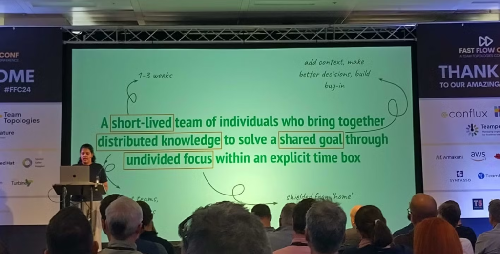 Varuna in front of a slide, which says: "A short-lived team of individuals who bring together distributed knowledge to solve a shared goal through undivided focus within an explicit time box"