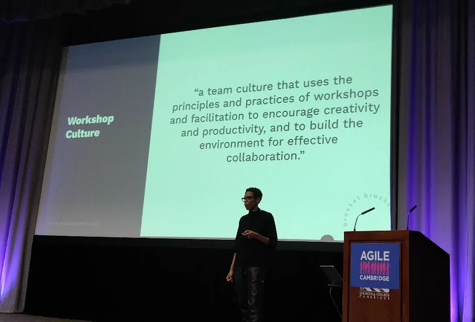 Alison on stage, in front of a huge slide defining workshop culture: "a team culture that uses the principles and practices of workshops and facilitation to encourage creativity and productivity, and to build the environment for effective collaboration."