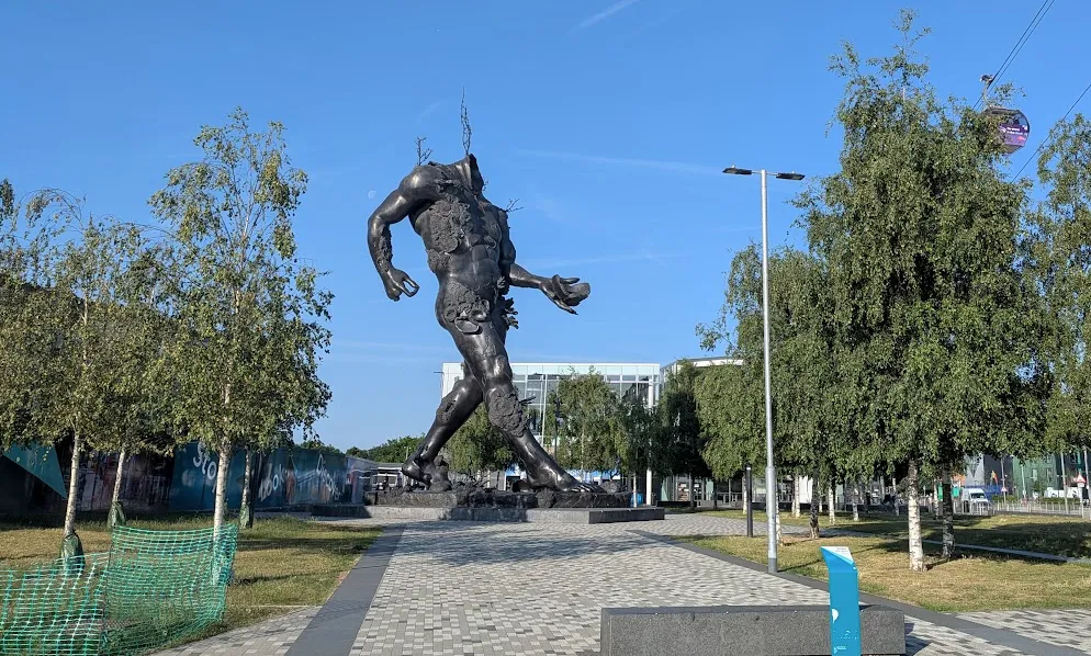 Photo of a huge metal headless figure, taller than the trees and buildings it seems to be walking past. It has no head, a broken bowl in its hand, and what looks like seaweed drifting upwards from various places.