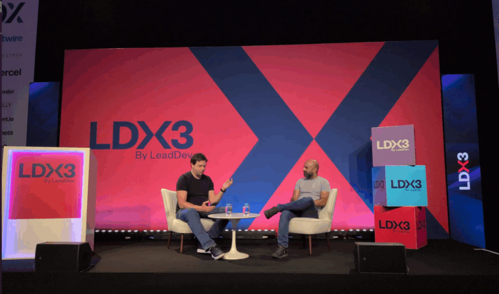 Gergely and Farhan sitting on chairs in front of a small table on a huge stage, with lots of conference logos around them.
