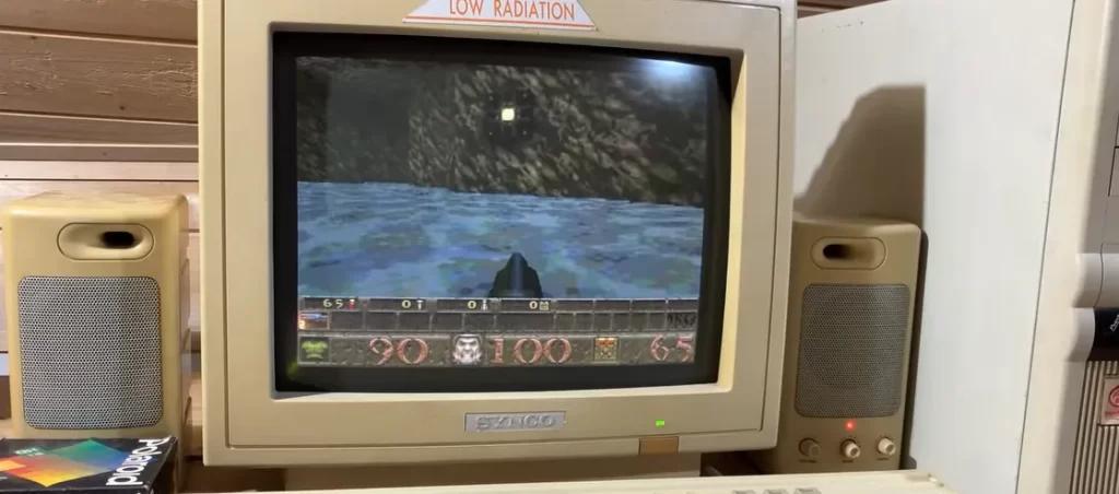 Screenshot from the video linked in the caption. Quake's a first person shooter game, the monitor shows a gun pointing forward from the bottom of the screen and a scene of water and rocks, with health and other stats at the bottom. It's an old-fashioned CRT monitor, with separate computer speakers, and a tall computer tower to one side, all very 1990s.