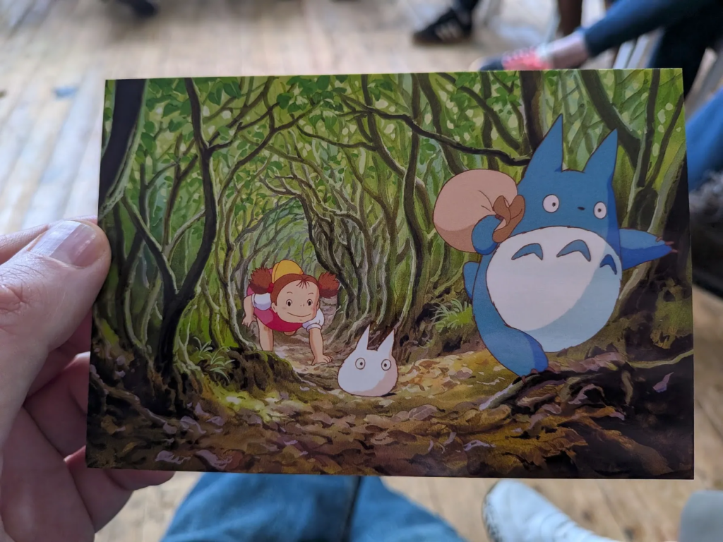 Hand holding a postcard with a scene from the movie My Neighbour Totoro. A cartoon of a small girl following 2 odd looking creatures through a tunnel of trees and branches.