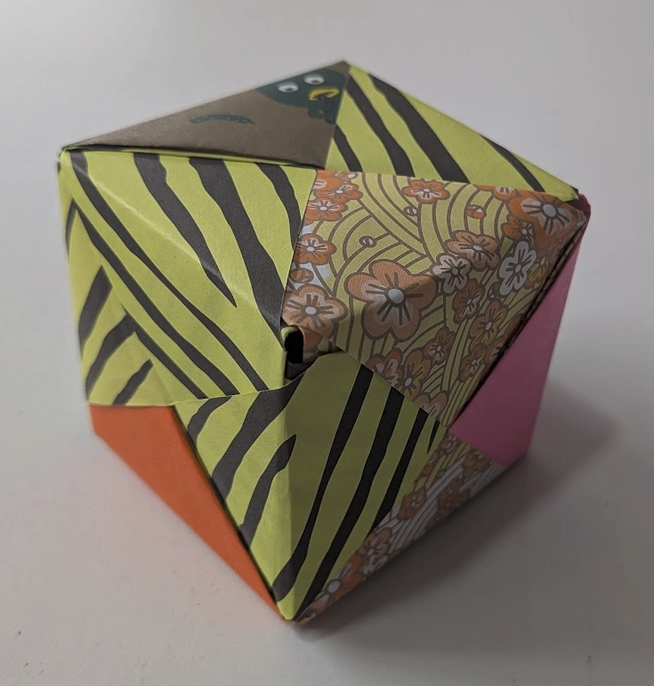 A cube made of folded origami paper. The various sheets of paper had different patterns, so the cube is a mismatched combination of solid colours, yellow and black tiger stripes, flowers, and a duck.