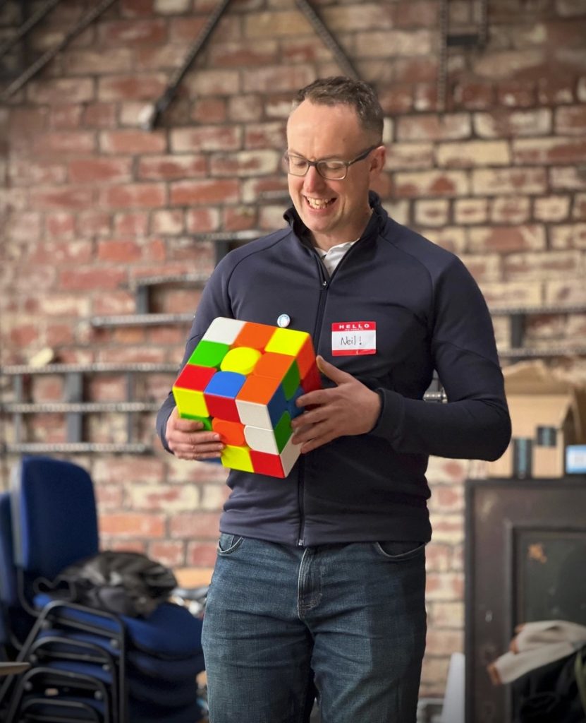 Photo of me holding a larger than usual cube (it's bigger than my head). I'm a 47 year old white man with short brown hair, glasses, wearing a zippy blue top and jeans.