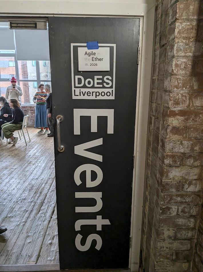 Photo of the door to the event, with a big "DoES Liverpool Events" sign printed on it and a piece of paper with "Agile in the Ether IRL 2026" stuck on there. It's a brick walled room with bare wooden floors. One side of the door is open and you can see people collaborating.