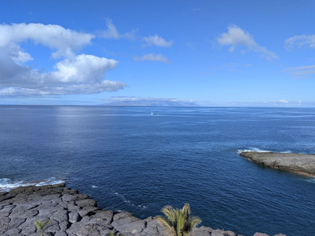 Photo of a calm bright blue sea, with a calm bright blue sky and a few clouds above it. In the foreground is a bit of rocky volcanic coast. In the distance are a few tiny boats, and an island on the horizon.