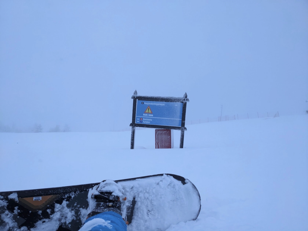 Photo of a snow-covered land with a cloud-filled sky, it's hard to tell which is which. In the foreground is a snowboard and a foot. In the middle of the frame there's a signpost pointing to various runs down the mountain. One is a black run with a large "steep" warning sign, the others are easier blue and red runs. All the writing is in Norwegian.