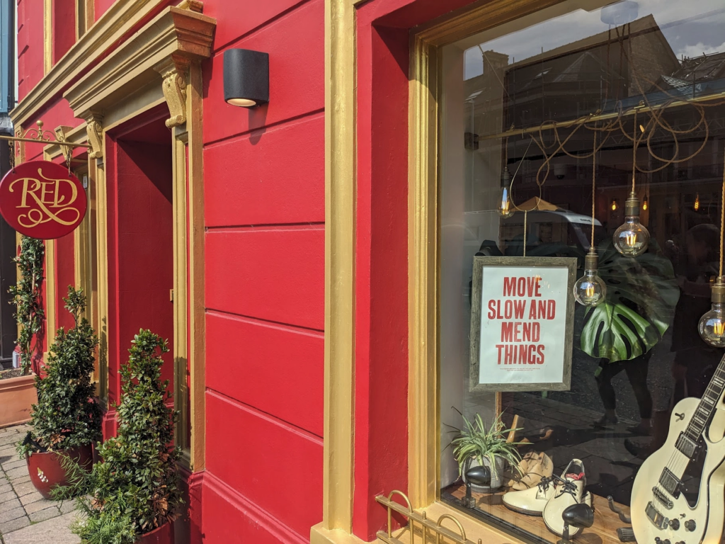 Photo of a shoemaker's shop: very dramatic red and gold outside, with a window that has a sign saying "Move slow and mend things".