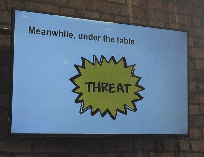 Slide saying "meanwhile, under the table" with a big comic-book style spiky yellow shape saying "threat".