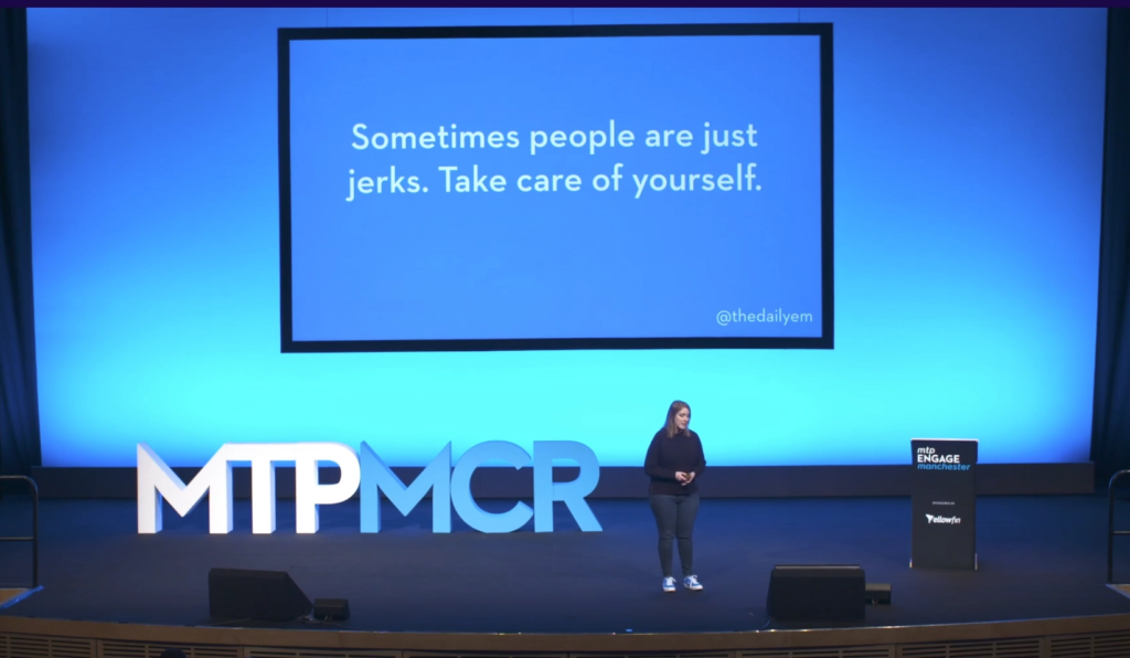 "Sometimes people are just jerks. Take care of yourself." is written in huge text on a plain slide blue slide. This is on a large screen behind Emily, who's on a stage at Mind the Product Engage in Manchester. 